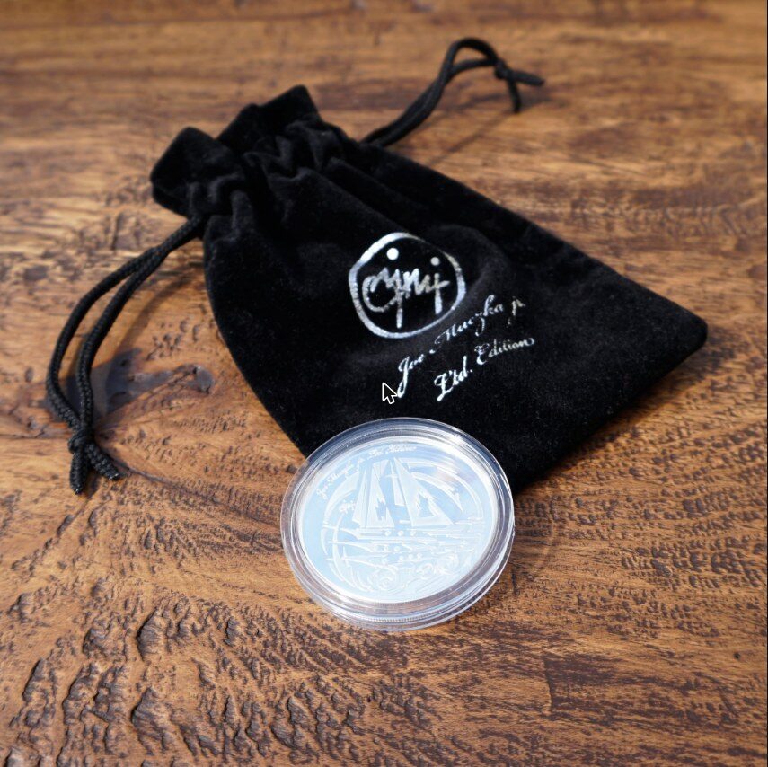Silver coin with a motif by Joe Muczka jr. in its capsule, next to the JMJ velvet pouch